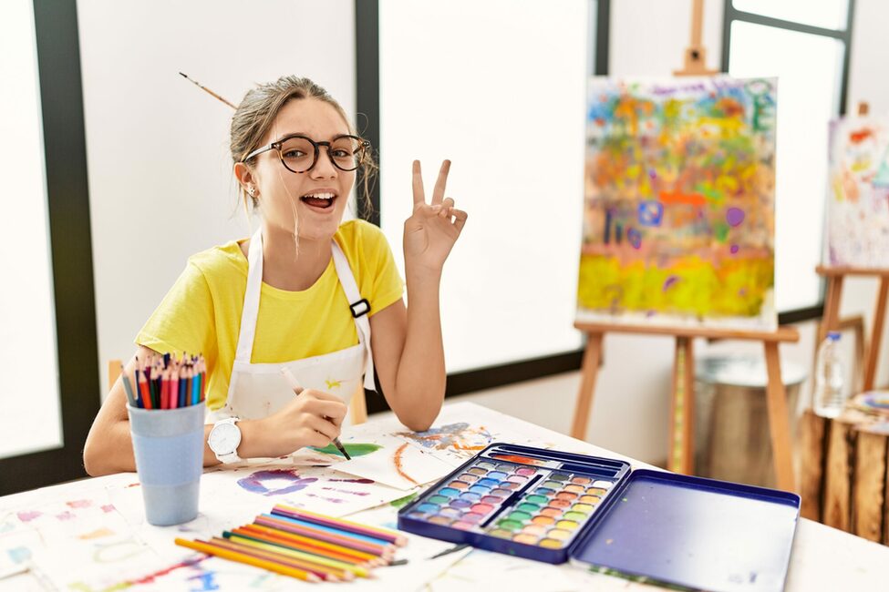 Ein Mädchen zeichnet mit bunten Stiften und macht ein Peace-Zeichen. Im Hintergrund steht eine bunte Leinwand.