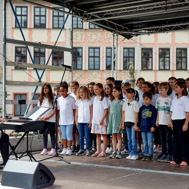 Eine Gruppe von Kindern steht auf der Bühne und singt ein Lied. Sie werden von einer Frau am Keyboard begleitet.