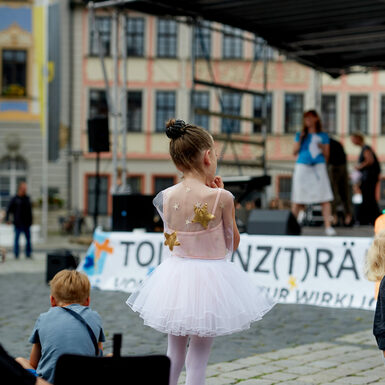 Ein Mädchen in Balletkleidung schaut zur Bühne, die im Hintergrund zu sehen ist.