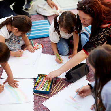 Kinder und eine Frau sitzen zusammen auf dem Boden und malen mit Buntstiften auf Papier.