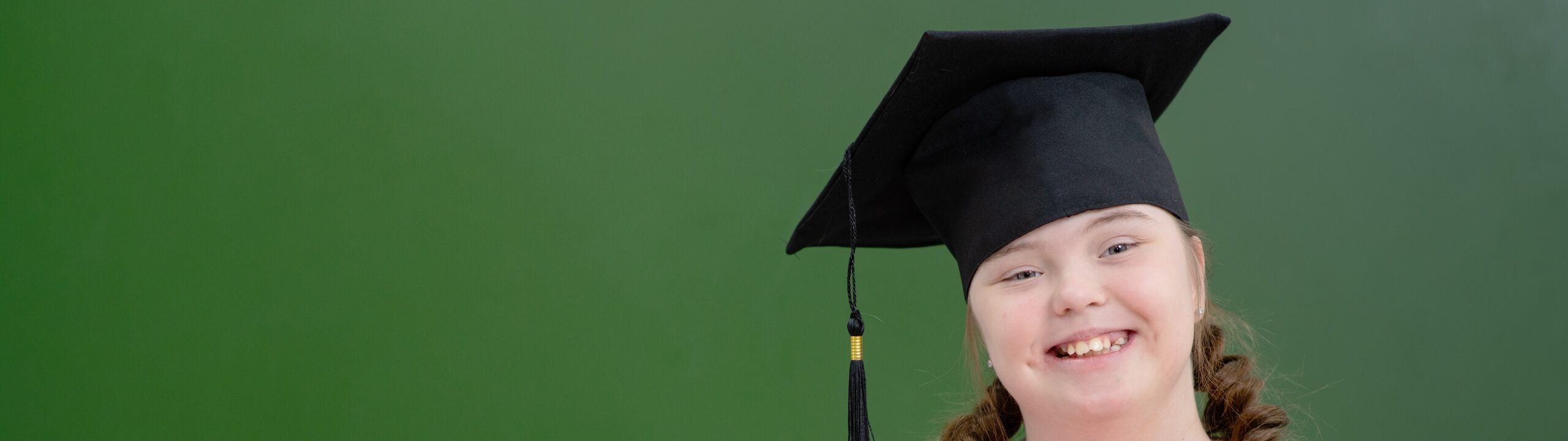 A teenager girl with Down syndrome wearing a graduate hat standing on the background of a green school board with a stack of books in her hands. Disabled girl with a smile looking at the camera